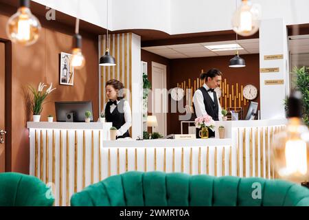 Professional employees checking reservations at front desk in hotel lobby to assist guests with check-in process. Two receptionists reviewing booking reservations waiting for arrival of visitors. Stock Photo