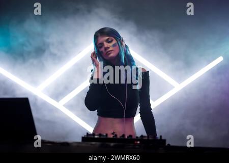 A female DJ with blue hair mixing music at a club, smoke and neon stage triangles in the background. Stock Photo