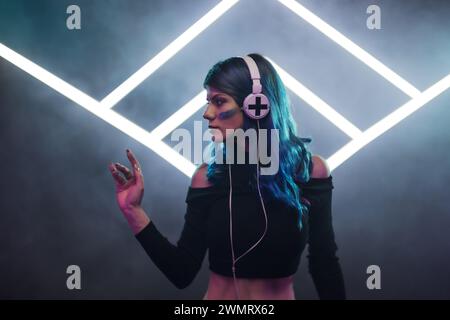 A female DJ with blue hair mixing music at a club, smoke and neon stage triangles in the background. Stock Photo