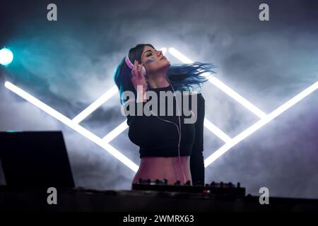 A female DJ with blue hair mixing music at a club, smoke and neon stage triangles in the background. Stock Photo