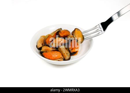 A fork picks up tinned canned smoked mussels from a dish, shellfish isolated on a white background. Stock Photo