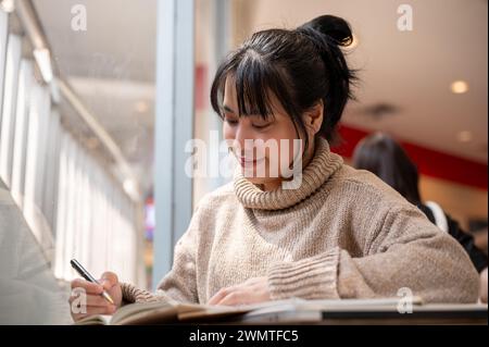 A beautiful and positive Asian woman in a comfy knitted sweater is keeping her diary or making a list of her ideas while working remotely at a cafe. Stock Photo