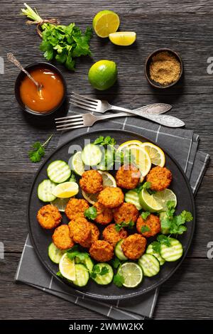 fried rice cutlets, rice tikki, indian style on black plate served with cucumber, lemon, lime slices and sweet chili sauce on dark wooden table, verti Stock Photo