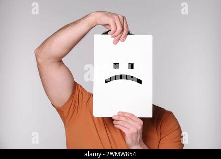 Man hiding behind sheet of paper with sad face on grey background Stock Photo