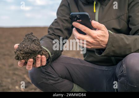 Agronomist is holding a clod of earth and smartphone, using the device to photograph the sample of the soil, selective focus Stock Photo