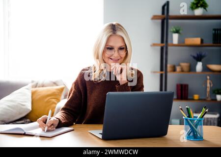 Photo of thoughtful middle age woman touch chin brainstorming writing notes in planner looking laptop during meeting session at office Stock Photo