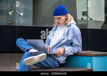 Long-haired stylish hipster guy sitting in street, looking at laptop screen, working as freelancer Stock Photo