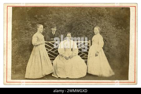 Original early to mid 1860's Victorian Carte de Visite (visiting card or CDV) group of Victorian women outside in the garden, lovely old bench, the older woman is seated on a bench, smiling boy in background wearing a bowler hat. Do not look like family members, so maybe servants, housekeeper or mistress seated. U.K.  circa 1865 Stock Photo