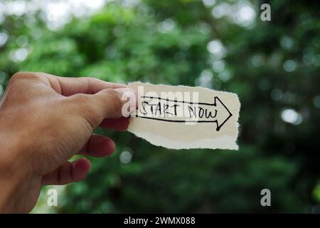 Hand holding a torn paper with the words 'Start now' on a tree branch, motivational typography. Stock Photo