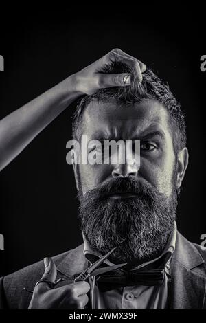 Bearded man hipster barber with beard and moustache in suit holds sharp scissors and cut hair in hairdresser salon. Barber scissors, barber shop Stock Photo