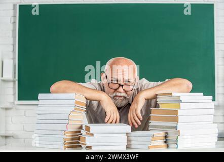 Teacher with too many books to read before exam. Teacher with books in school. Man school teacher standing in classroom. Old man teachers on green Stock Photo