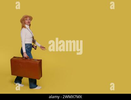 a woman in jeans, a white shirt, and  hat carrying a suitcase and taking photos by vintage camera on a yellow background. Happy people going on holida Stock Photo