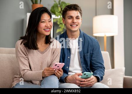 Diverse couple smiling and having fun using online phones. Joyful young adult couple using smartphone relaxing on a couch in their living room at home Stock Photo