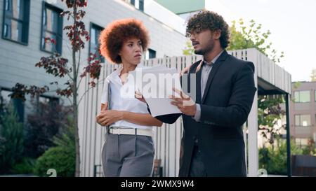 Indian Arabian male man businessman holding papers documents corporate discuss result plans with African American woman colleague businesswoman Stock Photo