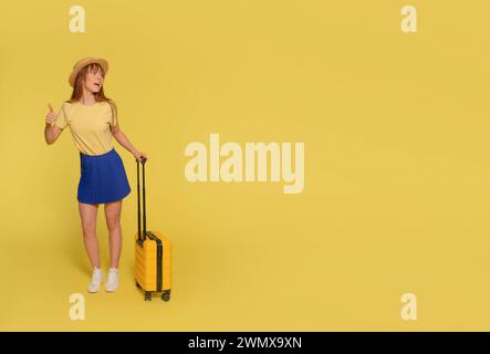 a happy young woman in blue skirt, a yellow T-shirt, and hat carrying a suitcase and give a thumb up on a yellow background. Happy people going on hol Stock Photo
