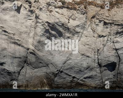 Weathered seaside rock face texture with parts of green and blue water. Aged volcanic stone wall surface background pattern with cracks and scratches. Stock Photo