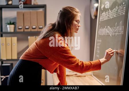 Side view medium shot of young Caucasian female music teacher writing on blackboard while preparing for lesson Stock Photo