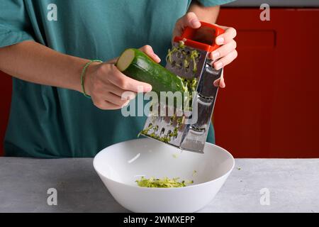 Authentic female hands grating green zucchini with a stainless steel hand grater into white bowl on kitchen table, close up, food lifestyle, vegan and Stock Photo