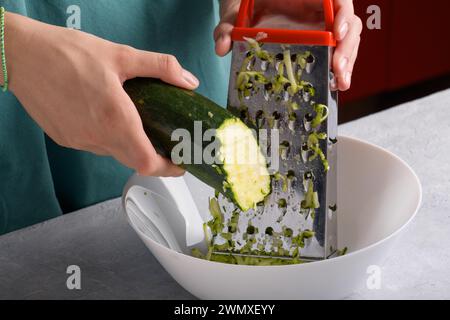 Authentic female hands grating green zucchini with a stainless steel hand grater into white bowl on kitchen table, close up, food lifestyle, vegan and Stock Photo