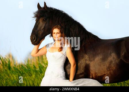 Friesian Horse. Jana Baade in a white dress standing with her gelding Nero in front of a reed belt. Germany Stock Photo