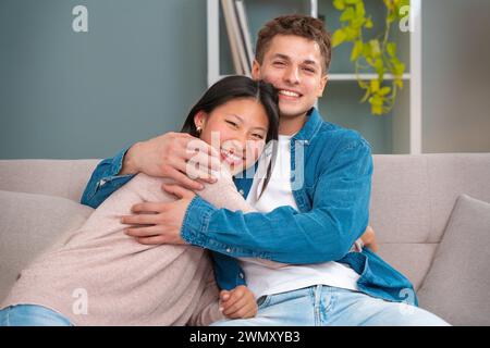Happy young multiracial couple hugging sitting on the sofa in the living room. Stock Photo