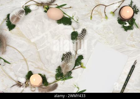 Handmade, Easter wreath on a marble table with white sheet of paper to fill. Eggs, eggshells, candles, moss - top view. Stock Photo