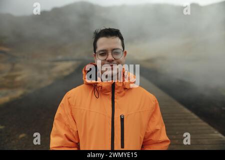 A smiling man in a bright orange jacket stands on a wooden boardwalk with a misty Icelandic landscape in the background. Stock Photo