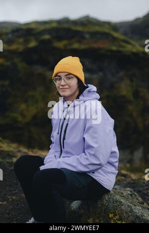A young woman wearing a yellow beanie and purple jacket is sitting on a rock with a moss-covered Icelandic hill in the background. Stock Photo