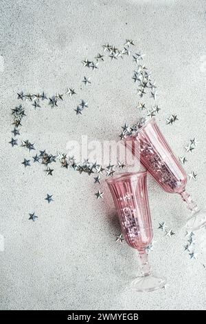 From above of two pink champagne glasses laid down with a trail of silver star-shaped confetti on a textured white surface Stock Photo