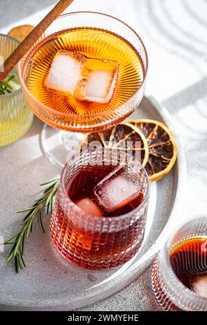 An array of exquisite alcoholic beverages in decorative glasses, accompanied by ice cubes, a cinnamon stick, rosemary, and dried citrus slices on a ma Stock Photo