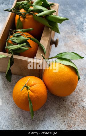 Top view of ripe oranges with fresh green leaves attached are displayed, some resting on a textured surface and others nestled in a wooden crate, evok Stock Photo