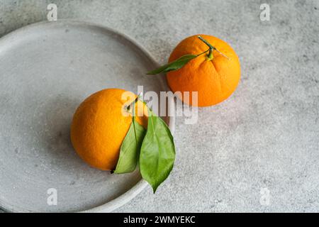 Two ripe oranges with vibrant green leaves, one in focus in the foreground on a concrete base with a blurred background Stock Photo