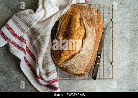 Top view of freshly baked loaf of rye sourdough bread sits cooling on a wire rack, accompanied by a bread knife and a white cloth with red stripes Stock Photo