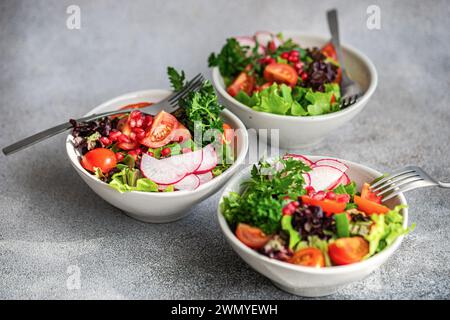 Three bowls of vibrant vegetable salad with lettuce, arugula, radishes, cherry tomatoes, green onions, and pomegranate seeds on a grey countertop Stock Photo