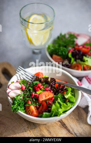 Top view of bowl of fresh vegetable salad with lettuce, arugula, radishes, cherry tomatoes, and pomegranate seeds on a wooden surface, beside a glass Stock Photo