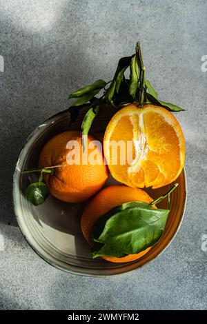 A ceramic plate holding fresh oranges, one sliced, with lush green leaves under natural light on a textured gray surface Stock Photo
