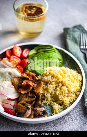 Close-up of a balanced lunch with a portion of bulgur wheat, a fried egg, avocado slices, cherry tomatoes, radishes, and brown mushrooms, accompanied Stock Photo