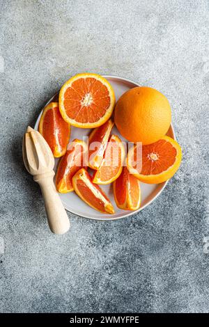 From above sliced and whole oranges arranged on a ceramic plate next to a wooden citrus juicer on a textured gray surface. Stock Photo