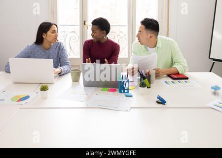 Three professionals actively engaged in a team discussion over documents at a workstation with laptops, colorful stationery, and data charts. Stock Photo