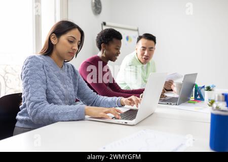 Three professionals collaborate using laptops in a well-lit workspace, exemplifying teamwork and diversity in a business environment. Stock Photo