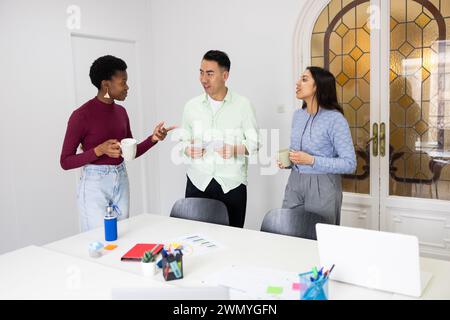 Three diverse professionals engaging in a lively discussion over a project document in a bright office setting, with cups of coffee in hand. Stock Photo