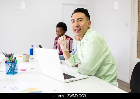 A smiling Asian man in a casual green shirt participates in an office meeting with colleagues, creating a vibrant and collaborative work atmosphere. Stock Photo
