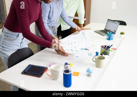 Two professionals are examining and discussing business charts and graphs on a white office desk, surrounded by office supplies. Stock Photo