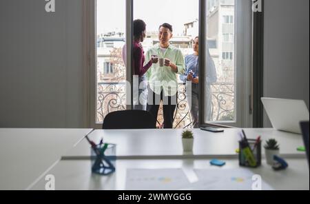 Three diverse colleagues chat and share a break by the window in a bright, open-plan office, with workstations visible in the foreground. Stock Photo