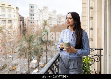 Young woman with a serene smile holding a coffee cup on a balcony with a cityscape backdrop. Stock Photo