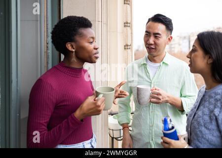 A group of diverse young adults engaged in a casual, friendly conversation while holding drinks on a balcony. Stock Photo
