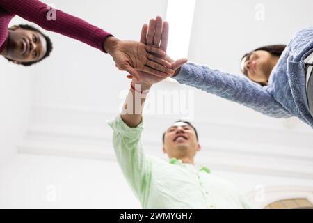 Diverse group of people stack hands together, signaling teamwork, unity, and collaboration in a light-filled room. Stock Photo