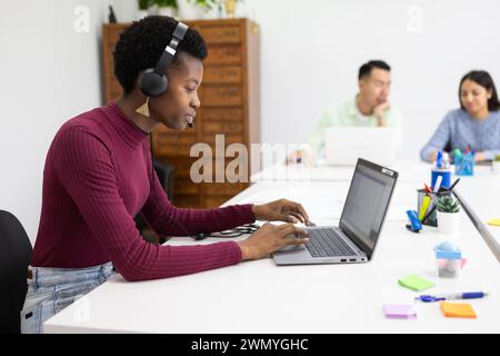 Young professional black woman working at her laptop with headphones in a modern office environment. Stock Photo