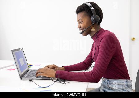 A cheerful customer service representative african female wearing a headset smiles while typing on a laptop at a modern office desk. Stock Photo