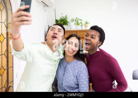 Three cheerful friends from diverse backgrounds share a moment of joy as they pose for a selfie with a smartphone in a bright indoor setting. Stock Photo